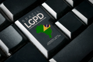 Imagem em close de um teclado preto, com destaque para uma tecla personalizada contendo a inscrição “Brasil LGPD - Lei Geral de Proteção de Dados Pessoais”. Na mesma tecla aparece a silhueta do mapa do Brasil acompanhada de elementos nas cores da bandeira nacional e o texto em inglês “General Data Protection Law”. O restante do teclado permanece desfocado ao redor, reforçando o foco na tecla relacionada à proteção de dados. No canto inferior esquerdo há um pequeno logotipo branco. A composição remete à segurança digital, privacidade de informações e conformidade com a legislação brasileira de proteção de dados.